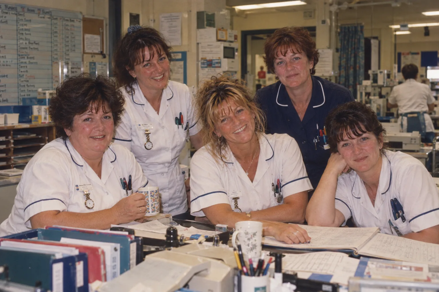 Five nurses at a busy ward desk in the 1990s, Susan in the middle with her sleeves rolled up, all smiling at the camera.
